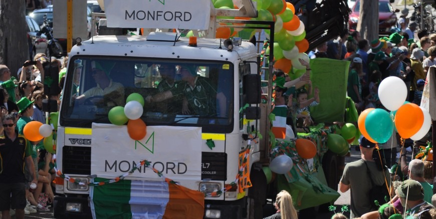 A photograph of a Monford truck during a a parade with streamers, balloon, and flags hanging of it. The families of employees waving to the crowds from the back.