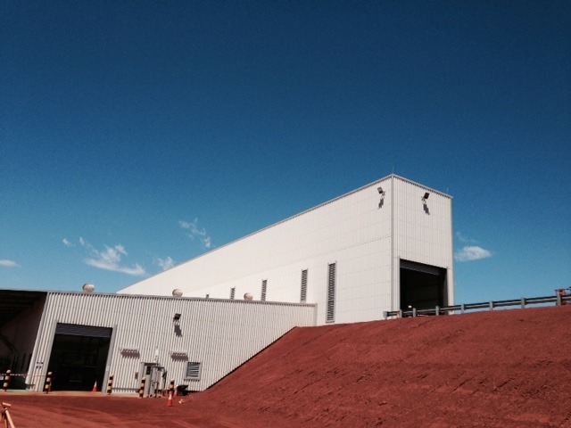 This photograph shows a large warehouse used at Nammuldi Mine. It is surrounded by red dirt at bright blue sky.